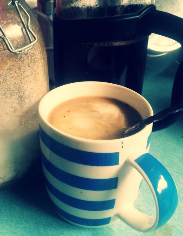 [food and drink] Coffee with coconut cream. [image description: On a light-blue benchtop sits a fairly fat cup with coffee in it. The coffee has been whitened with coconut cream, which has left two bright patches on the surface of the coffee. Behind t